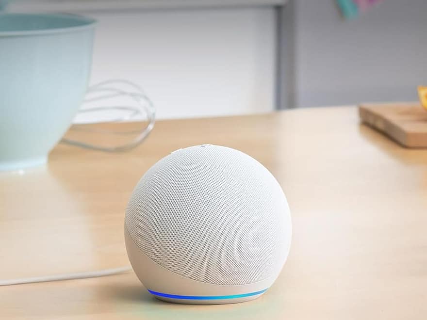 Echo Dot (newest model) spherical fabric smart speaker in white with blue light ring on wooden countertop in kitchen setting
