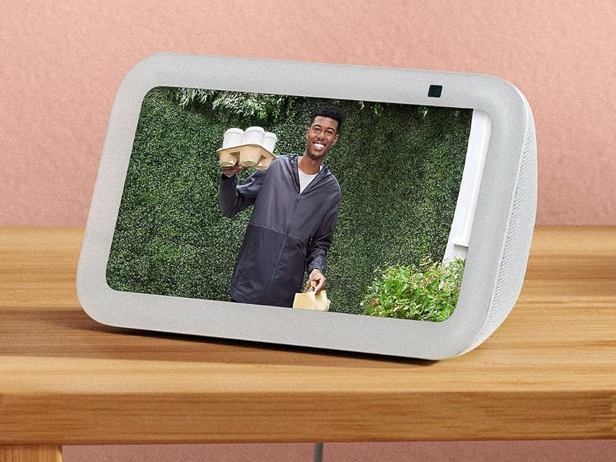Amazon Echo Show 5 (newest model) smart display in Glacier White with angled fabric base on table, screen showing Alexa door camera view