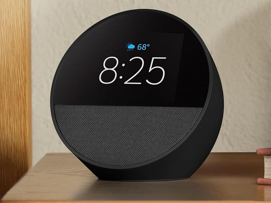 Amazon Echo Spot (newest model) black round smart alarm clock with display showing time, fabric speaker base on wooden nightstand