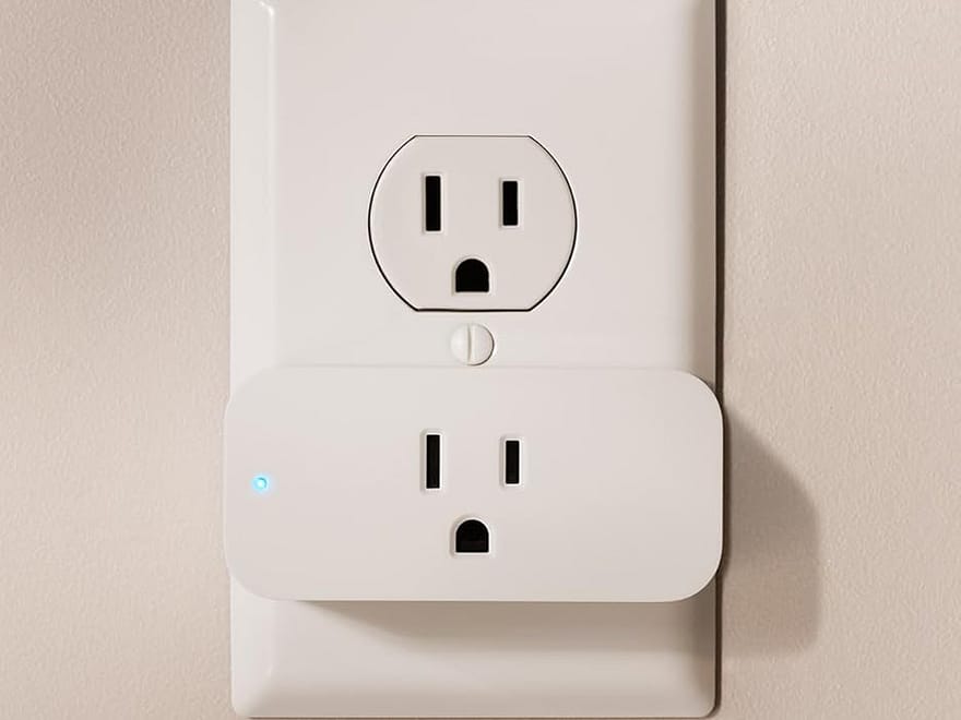 Amazon Smart Plug in a white wall outlet, rectangular white finish with blue indicator light and Alexa use icons below